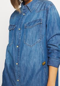 Camicia in denim con colletto, maniche lunghe, due tasche frontali, bottoni dorati e un'etichetta con il logo G-Star sul lato. Tessuto blu e strutturato.