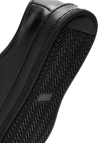 Zapatilla negra que muestra una suela de goma texturizada con un patrón de zigzag, parte superior de cuero suave y costuras visibles a lo largo de los bordes.
