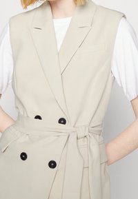 Beige ärmlös väst i slätt tyg, med dubbelknäppt design, svarta knappar och ett knytbart midjebälte för en figursydd look.