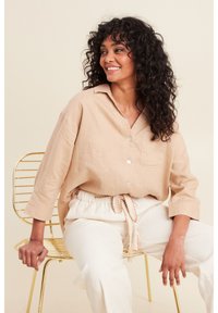Camicia button-up in lino beige con colletto e tasca, abbinata a pantaloni bianchi con coulisse. Sfondo color crema e sedia in filo dorato.