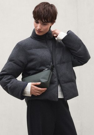 Persoon draagt een donkergrijze donsjas en zwarte broek en houdt een kleine donkergroene crossbodytas vast tegen een effen lichte achtergrond.