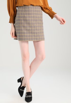 Donna con minigonna marrone a quadri, camicetta lunga color ruggine e scarpe nere con tacco con decorazioni.