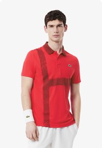 Röd pikétröja med en brun krage, som har ett mörkt abstrakt mönster. Modellen bär även ett vitt handledband med en grön logotyp.