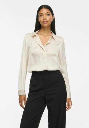 Femme debout portant un chemisier en soie crème à manches longues rentré dans un pantalon noir taille haute sur un fond uni.