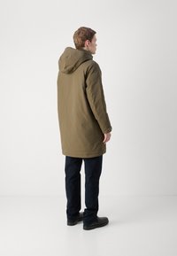Olivgrüner Parka mit Kapuze, verfügt über eine glatte, matte Oberfläche und einen etwas längeren Schnitt. Kombiniert mit dunklen Hosen und schwarzen Schuhen.