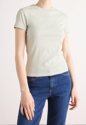 Person wearing a fitted light beige short-sleeve t-shirt and blue denim jeans, standing against a plain light background.