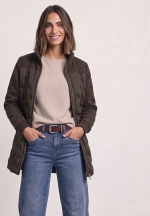 Femme souriante aux cheveux bruns mi-longs, portant une veste matelassée foncée ouverte, un pull beige, un jean bleu et une ceinture marron, mains dans les poches.
