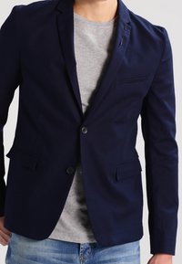 Navy blazer with two buttons, a single chest pocket, and two front pockets; paired with a grey t-shirt and blue denim jeans.