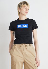 Juodas trumpomis rankovėmis marškinėliai su dideliu mėlynu stačiakampiu logotipu, kuriame užrašyta "HUGO" baltomis raidėmis, derinami su smėlio spalvos šortais su raišteliu.