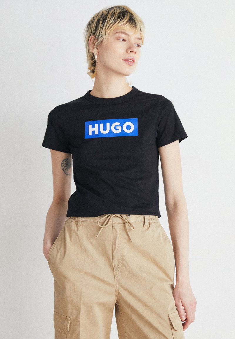 Juodas trumpomis rankovėmis marškinėliai su dideliu mėlynu stačiakampiu logotipu, kuriame užrašyta "HUGO" baltomis raidėmis, derinami su smėlio spalvos šortais su raišteliu.