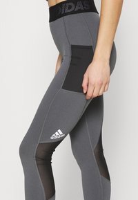Leggings sportivi grigio scuro con vita alta, dotati di pannelli in mesh neri e logo Adidas bianco sulla parte inferiore della gamba. Tessuto liscio.