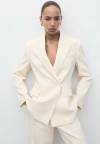 Lichtbeige op maat gemaakte blazer met een diepe V-hals, sluiting met één knoop en twee voorkhockets. Gladde stof en gestructureerd ontwerp.