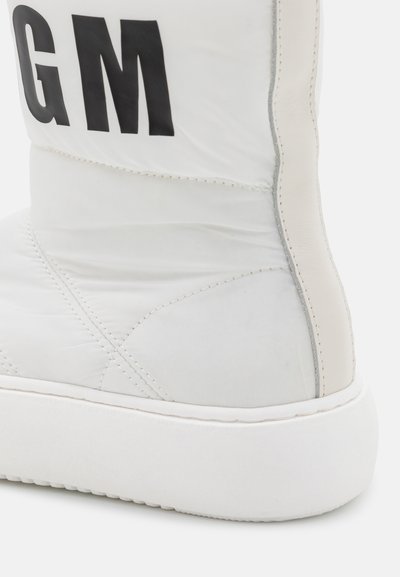 Bota acolchoada branca com um cabedal sintético liso, apresentando letras pretas "GM", sola branca e detalhes em costura ao longo das laterais.