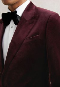 Giacca in velluto bordeaux con revers a punta, taschino e papillon nero. Camicia bianca con texture e bottoni neri visibili.