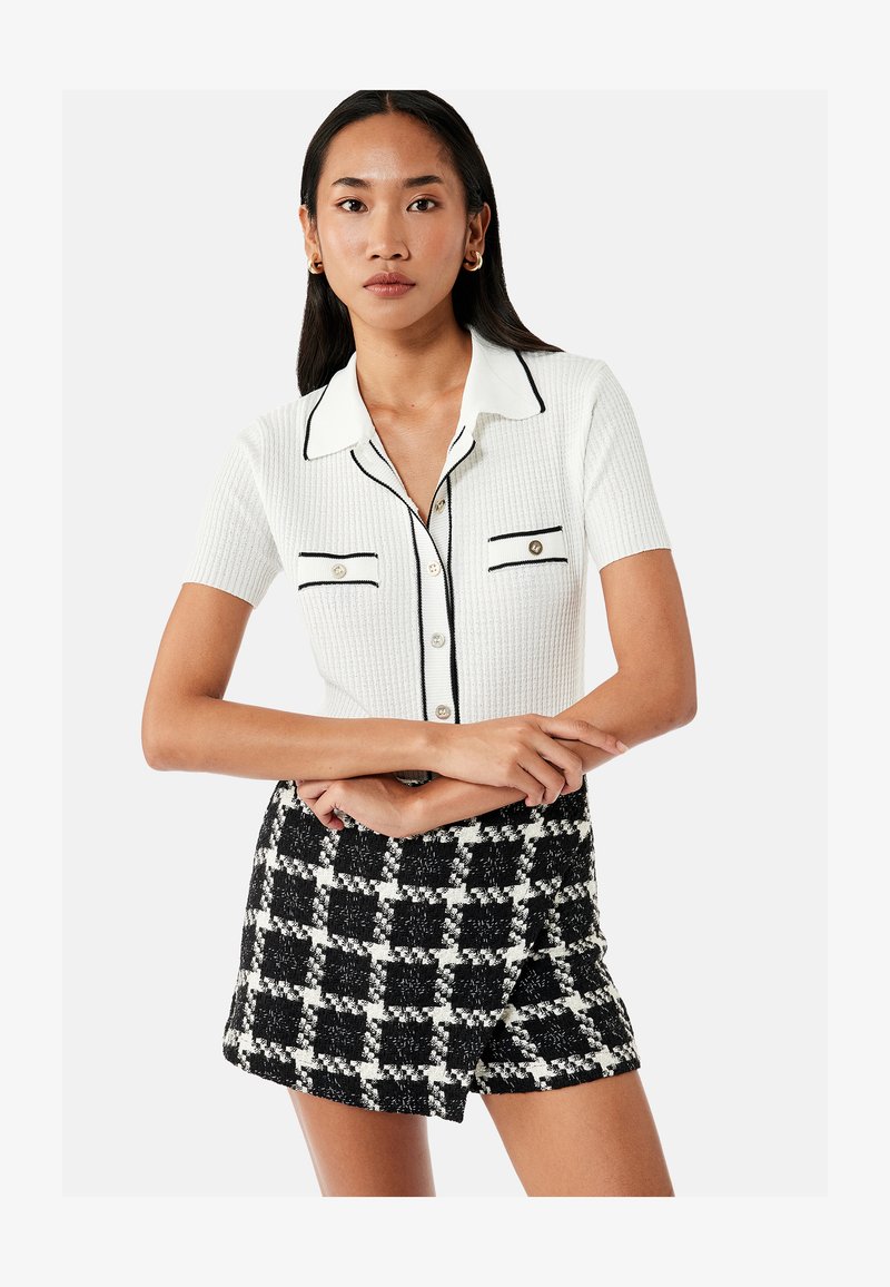 Junge Frau steht mit verschränkten Armen, trägt ein weißes kurzärmeliges Hemd mit schwarzem Besatz und schwarz-weiß karierte Shorts.