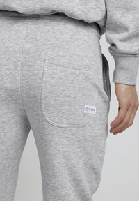 Lichtgrijze trainingbroek met een achterzak, zachte textuur, elastische tailleband en een klein merklabel aan de linkerbenedenkant.