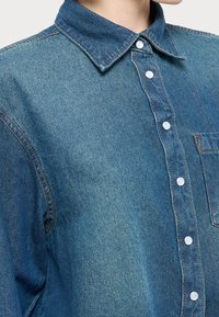 Camicia di denim con colletto a punta, tasca frontale e chiusura con bottoni bianchi. Il tessuto è di un blu medio con una texture scolorita.