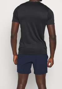 Vista trasera de una persona con una camiseta negra de manga corta y pantalones cortos azul marino contra un fondo liso.