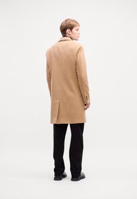 Camel-colored coat with a straight cut, notch lapels, single vent, and buttoned cuffs. Worn over black trousers and shoes.
