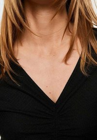 Černý top s výstřihem do V a řasením na výstřihu. Hladká textura látky kontrastuje s tónem pleti a vlasy nositele.