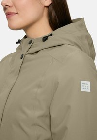 Close-up van een persoon die een beige hooded jas draagt met zwarte knopen en een "NEW VIE W<<" logo op de linkermouw.