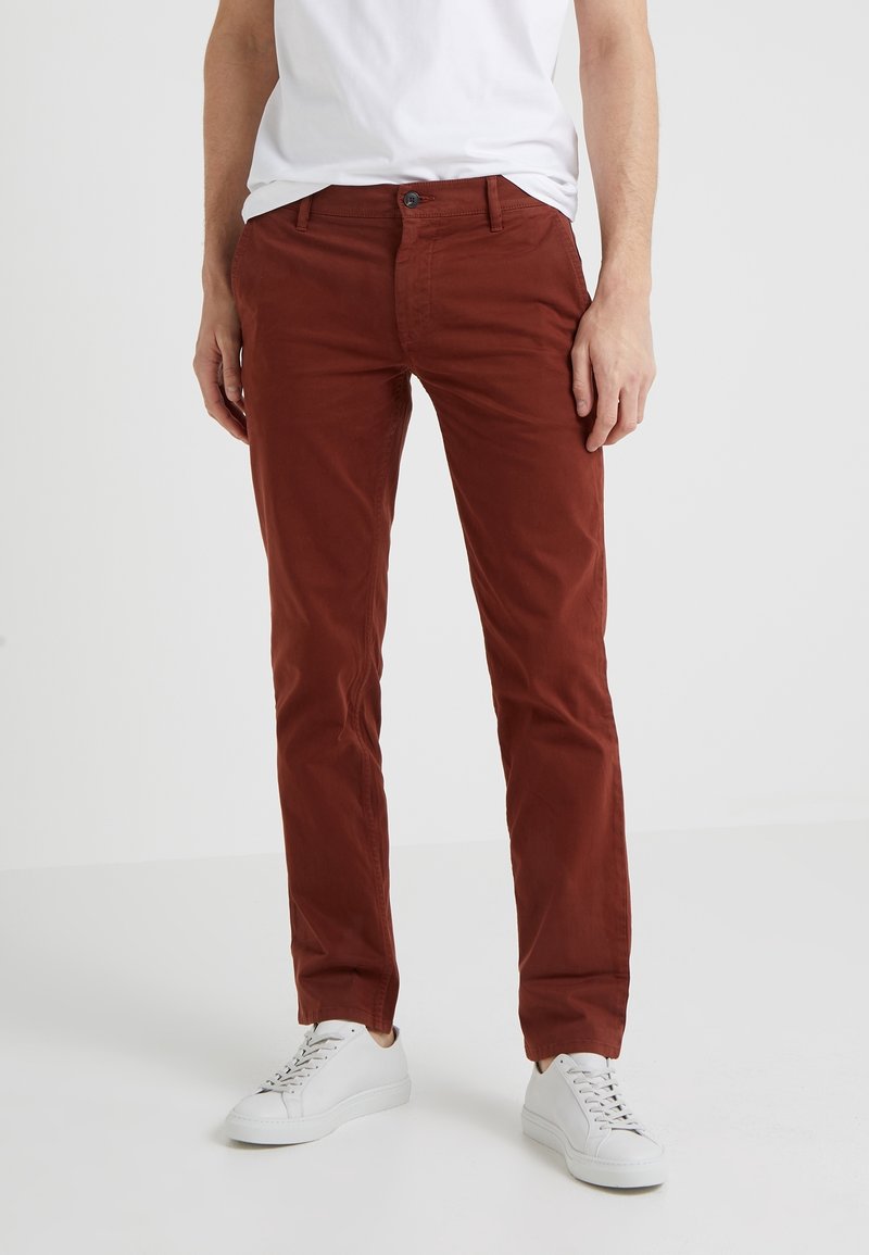 Hnědé bavlněné chino kalhoty s úzkým střihem, s knoflíkovým zapínáním a pěti kapsami. Kombinovány s bílými sportovními botami.