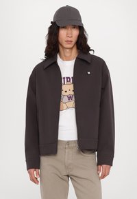 Jachetă zip-up de culoare marron închis, cu guler ascuțit și patch cu logo, purtată peste un tricou alb cu grafică și pantaloni bej, completată cu o șapcă gri.