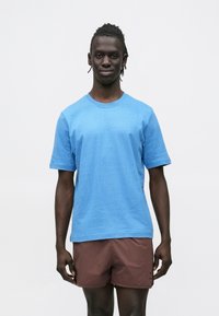 Hele sinine puuvillane T-särk lühikeste varrukatega ja ümar kaelusega, kombineeritud pruunide lühikeste pükstega. Sile tekstuur ja mugav istuvus.