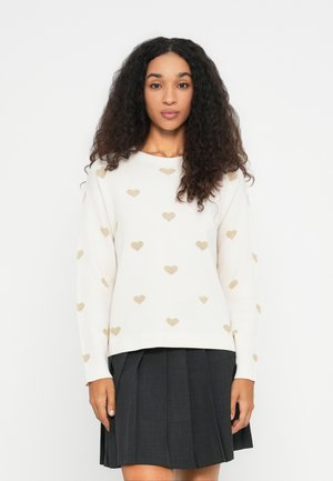 Femme avec de longs cheveux bouclés portant un pull blanc avec des cœurs dorés et une jupe plissée noire, se tenant devant un fond uni.
