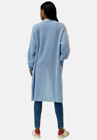 Cardigan lavorato a maglia azzurro chiaro con maniche lunghe e vestibilità ampia. Design con schiena scoperta, polsini a coste e tasche laterali. Abbinato a jeans e sneakers.
