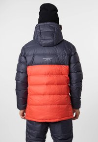 Puffermantel mit oberem Teil in Marineblau und unterem Teil in leuchtendem Rot, gesteppter Design, mit Kapuze, aus leichtem Material, mit sichtbarem Branding auf der Rückseite.