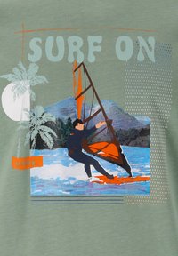 Vihreä T-paita, jossa on graafinen design tuulenpurjehtijasta, oranssista purjeesta, palmuista ja rohkeasti kirjoitetut sanat "SURF ON".