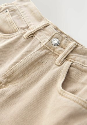 Beige Denim-Shorts mit einem vorderen Metallknopfverschluss, sichtbaren Nähten und einer Taschenapplikation. Das Material hat eine glatte, leicht strukturierte Oberfläche.