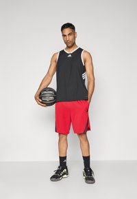 Mand i sort ærmeløs sports top, røde shorts og sorte sneakers holder en sort Spalding basketball mod en ensfarvet baggrund.
