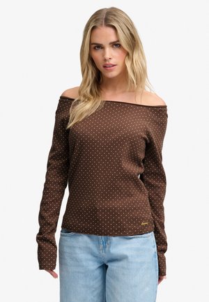 Lyshåret kvinde iført brun skjorte med lange ærmer uden skuldre med hvide polkaprikker og lyseblå jeans, står mod en ensartet baggrund.