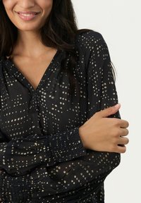 Blusa scura e trasparente con pois dorati. Presenta un scollo a V e maniche con polsini arricciati. Tessuto morbido e leggero con una texture liscia.