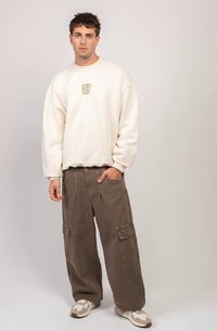 Jovem em pé com uma mão no bolso, vestindo um sweatshirt oversized em creme, calças cargo marrons largas e sapatilhas brancas contra um fundo simples.