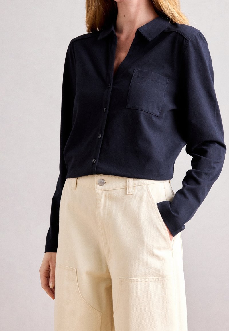 Femme portant une chemise boutonnée bleu marine foncé à manches longues avec une poche poitrine et un pantalon beige taille haute, main dans la poche.