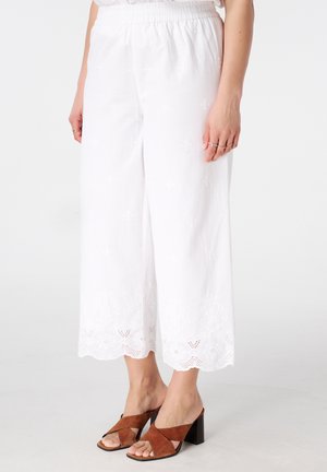 Vrouw die witte, wijd uitlopende, korte broek draagt met geborduurde geschulpte zoom en bruine sandalen met blokhak en open teen.