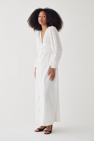 Kvinde med krøllet hår iført en lang, hvid kjole med hulmønster, puffede ærmer og sorte åbne sko med hæl mod en ensartet, lys baggrund.