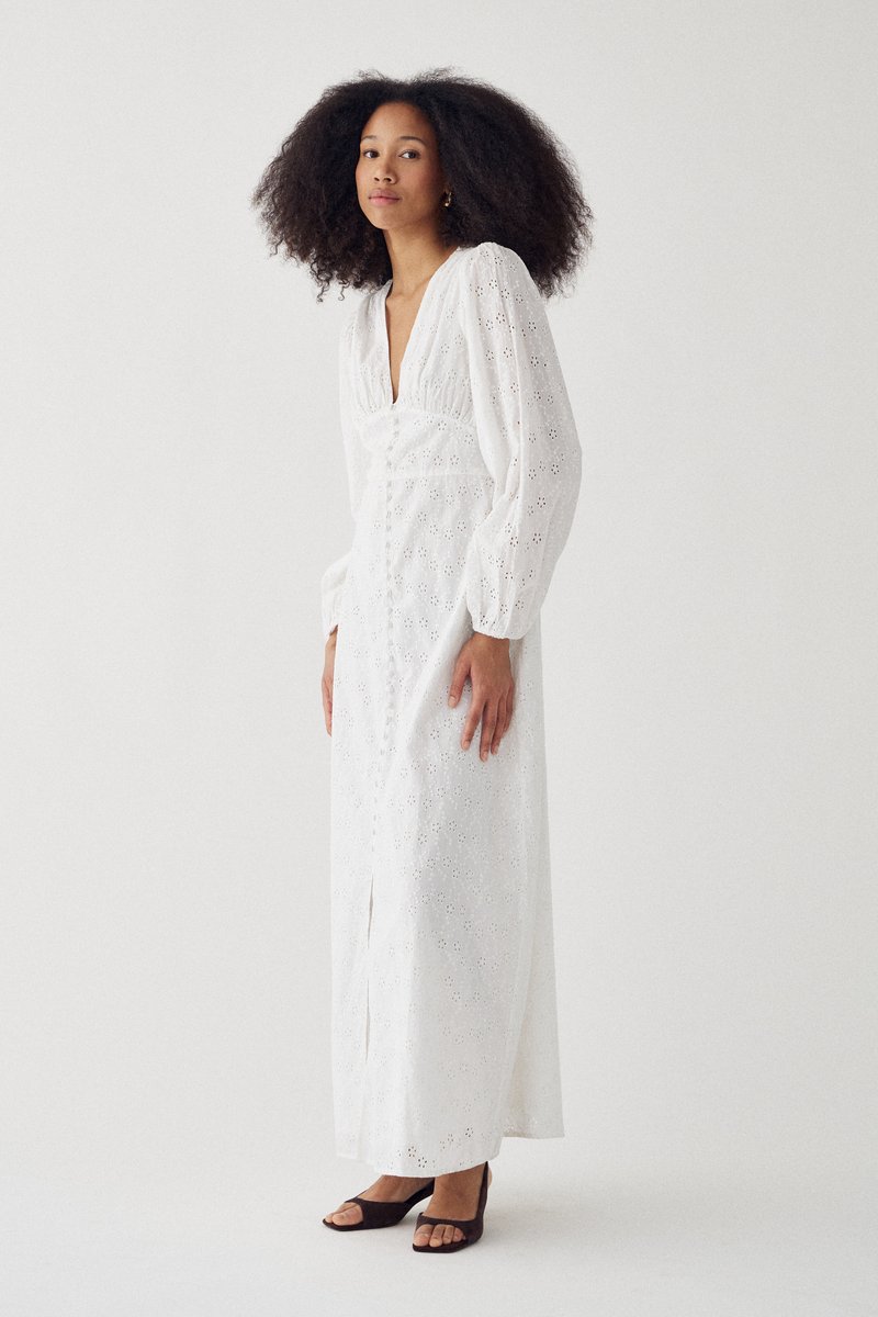 Vrouw met krullend haar, gekleed in een lange witte oogjesjurk met pofmouwen en zwarte open-toe hakken, tegen een effen lichte achtergrond.