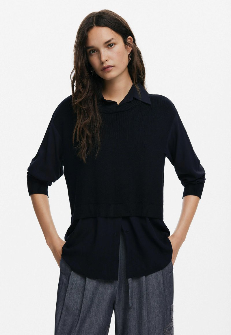 Schwarzer Strickpullover mit einem runden Ausschnitt, über einem dunklen Hemd mit Knöpfen getragen, kombiniert mit grauen, gefalteten Hosen.