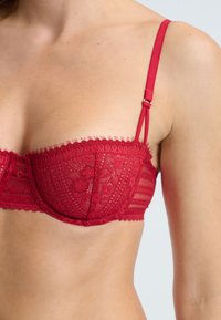 Reggiseno rosso in pizzo con un design strutturato, bordi ondulati e spalline regolabili. Presenta motivi floreali e una finitura satinata liscia.