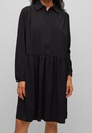Black long-sleeve button-up dress with a gathered waist and collar worn by a person against a plain gray background.