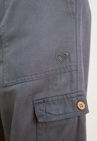 Pantalon cargo en coton gris avec une poche latérale munie d'un bouton en bois et un logo brodé sur la cuisse. Tissu texturé avec une brillance subtile.