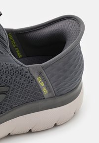 Grå slip-on sneaker med mesh-övre, texturerade detaljer och en tjock vit sula med ett mönstrat mönster. "SKECHERS SLIP-INS"-etikett synlig.
