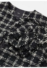 Veste en tweed texturée noir et blanc avec un motif à carreaux et un détail plissé près de l'encolure, étiquetée « MNG ».