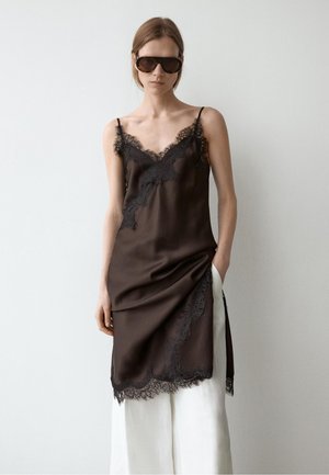 Kvinde iført store mørke solbriller og en sort blondetrimmet slipkjole lagdelt over hvide vidde bukser, stående op ad en plain væg.