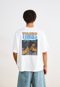 Camiseta blanca de manga corta con un diseño gráfico de botas marrones sobre un fondo negro y azul, con el texto "TIMBS ON MY FEET".