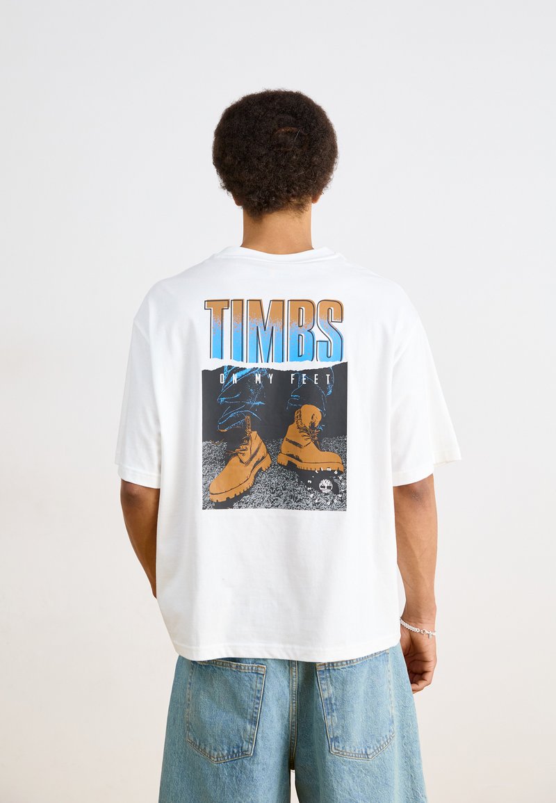Camiseta blanca de manga corta con un diseño gráfico de botas marrones sobre un fondo negro y azul, con el texto "TIMBS ON MY FEET".
